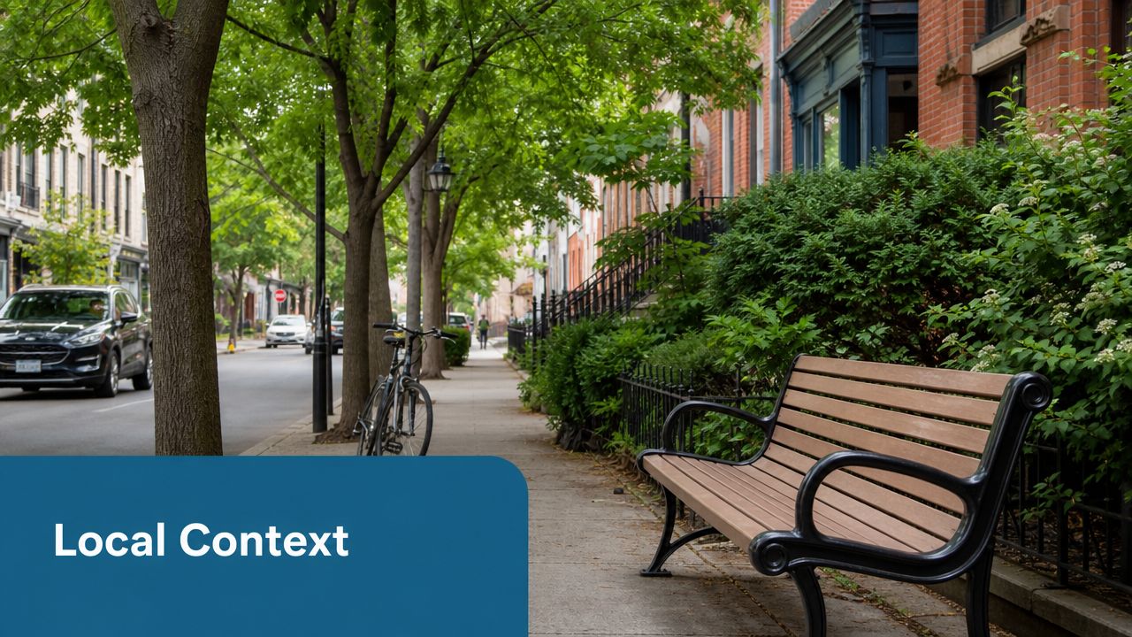 A quiet residential street lined with green trees, a park bench, and brick houses in the city.